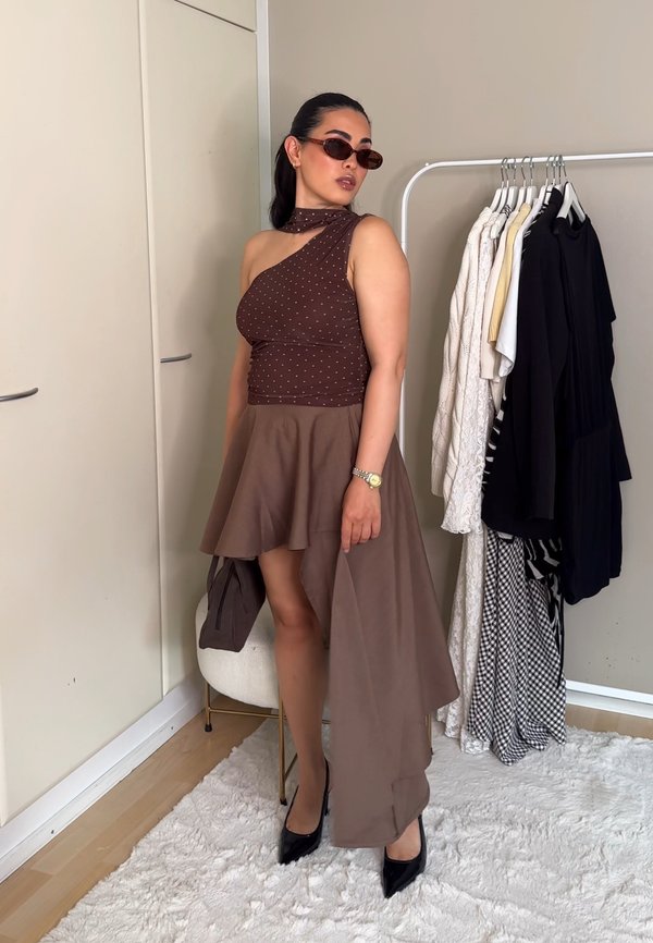 Mujer con vestido marrón asimétrico y gafas de sol, de pie junto a un armario con varias prendas colgadas en una habitación luminosa.
