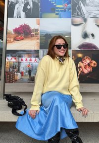 Pull jaune clair, jupe en satin bleue, bottes noires montantes jusqu'aux genoux. En arrière-plan, plusieurs photographies et un sac à bandoulière noir à proximité.