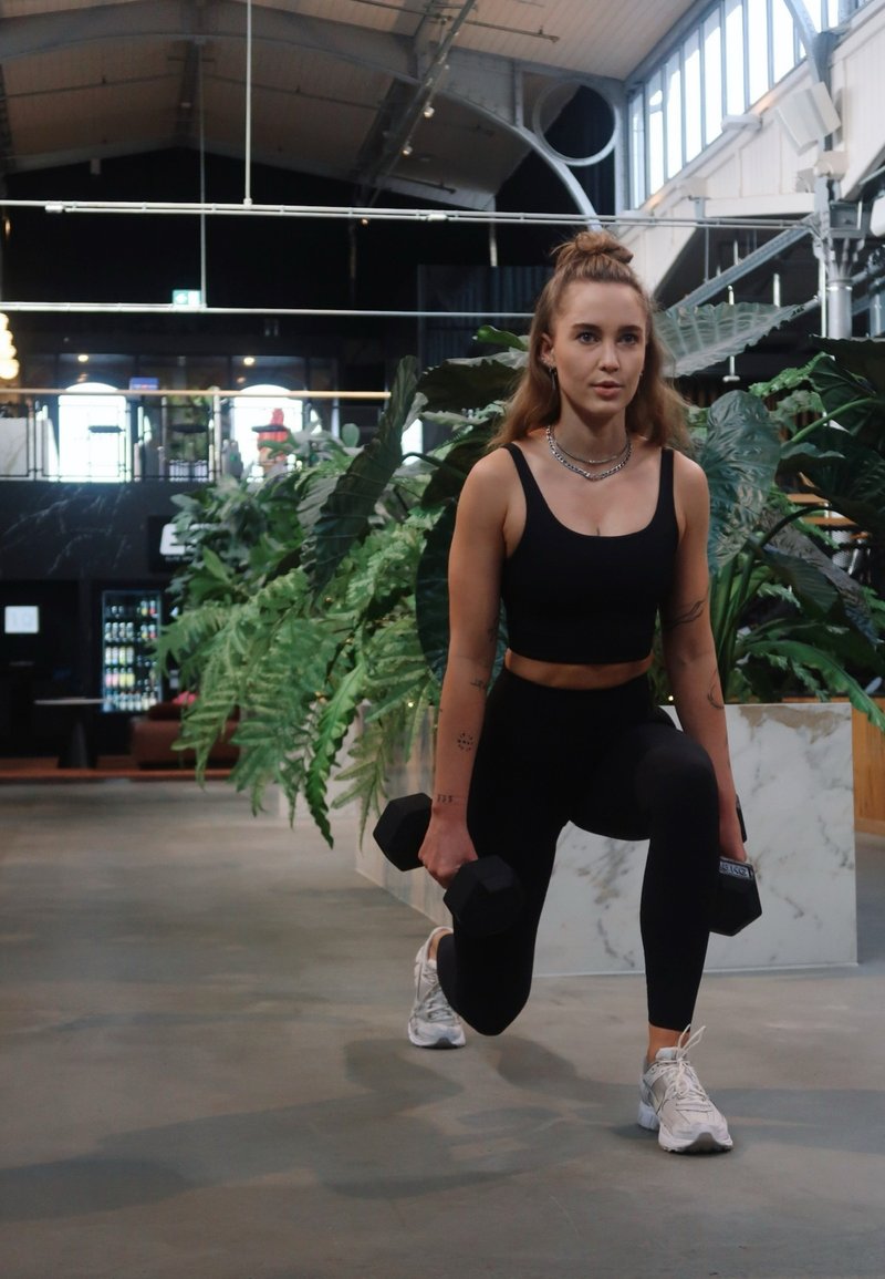 Top atletico nero e leggings; donna che esegue un affondo tenendo manubri neri; scarpe da ginnastica bianche; sfondo con vegetazione.