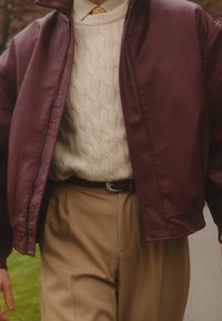 Blouson aviateur en cuir marron foncé porté sur un pull en maille torsadée crème, associé à un pantalon beige et une ceinture marron. Design texturé et sur mesure.