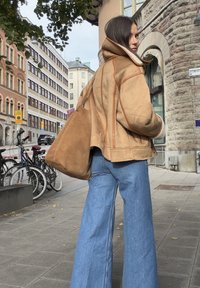 Beige mokkasinko takki oranssilla ompeleilla, yhdistettynä vaaleansinisiin leveälahkeisiin farkkuihin. Ruskea olkalaukku. Kaupunkikatuympäristö, jossa on polkupyöriä.