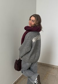 Jeune femme en pull gris et jean avec une écharpe et un sac bordeaux, regardant par-dessus son épaule, debout à l'intérieur sur un sol carrelé.