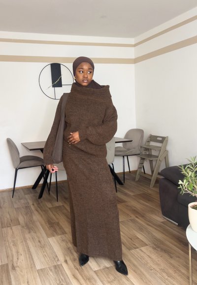 Suéter marrón de punto grande con cuello alto y mangas largas, combinado con una falda larga a juego. Tacones negros puntiagudos. Interior moderno.