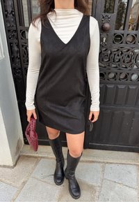Rochie neagră din suede artificial, cu decolteu în V, purtată peste un top crem cu mâneci lungi. Asortată cu cizme negre până la genunchi și o geantă burgundă.