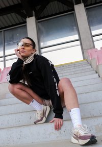 Sweat-shirt noir à manches longues avec imprimé blanc, short beige, chaussettes blanches aux chevilles, et baskets crème/violettes. Grandes lunettes de soleil teintées. Escaliers en béton.