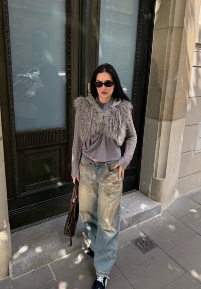 Chaqueta corta gris de felpa sobre una blusa gris, combinada con jeans de pierna ancha de un azul claro. Lleva zapatos negros y sostiene un bolso marrón. Gafas de sol negras.