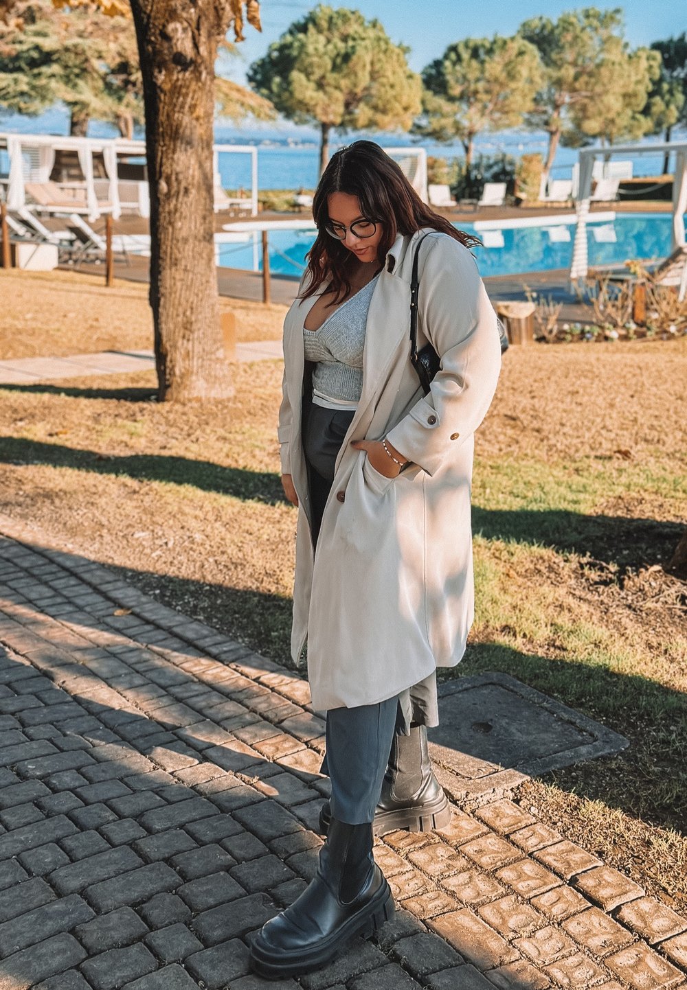 Uma mulher com um trench coat bege claro sobre um top cinza e umas calças escuras está em um caminho de paralelepípedos, usando botas de tornozelo pretas.