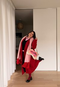 Femme en manteau rouge, écharpe rose avec franges, et chaussures à plateforme noires posant une jambe levée à l'intérieur près de rideaux blancs et d'un sol en bois.