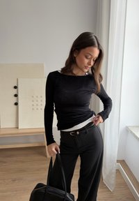Jeune femme portant un haut à manches longues noir et un pantalon, tenant un sac noir, debout à l'intérieur près d'une fenêtre avec des rideaux légers et des panneaux d'art abstrait.