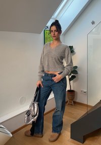 Femme aux cheveux foncés attachés en chignon, portant un pull gris court et un jean bleu, tenant un sac argenté, debout dans une pièce lumineuse près d'une plante.