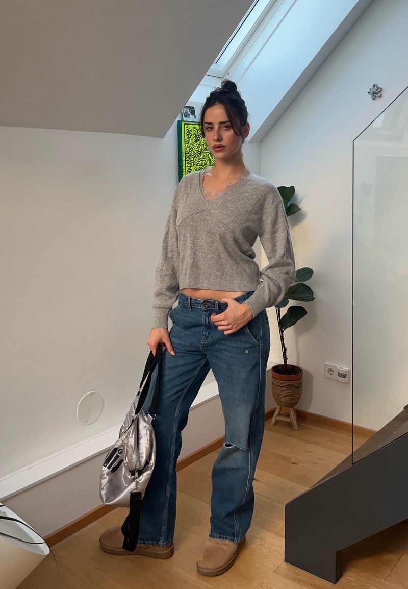 Frau mit dunkel gebundenem Haar trägt einen grauen cropped Pullover und blaue Jeans, hält eine silberne Tasche und steht in einem hellen Raum neben einer Pflanze.