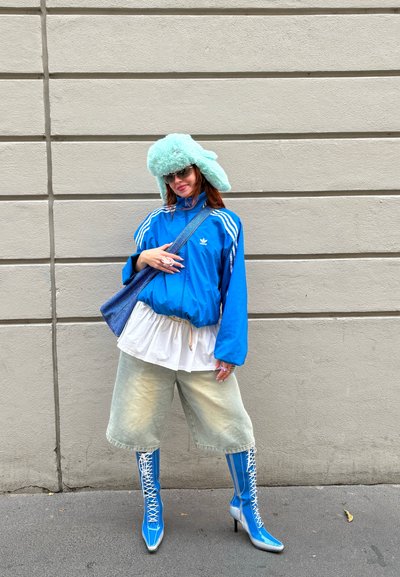 Rompeviento azul de Adidas con rayas blancas, camiseta blanca oversized, pantalones cortos verdes claros y botas altas azules con cordones. Un gorro de felpa en tono teal añade textura.