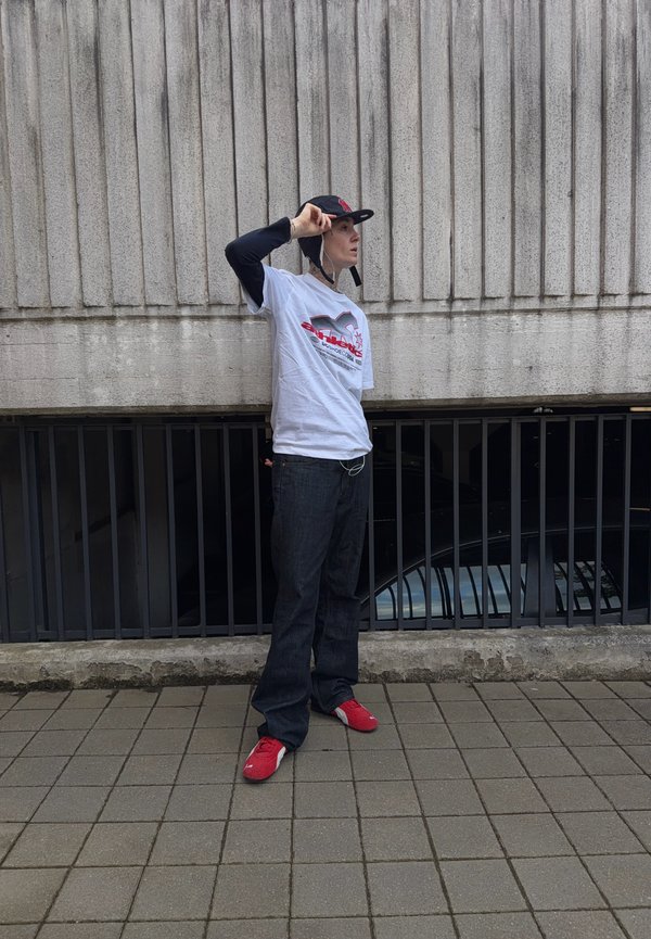 Personne portant un t-shirt blanc à motif, des manches longues noires, une casquette noire, un pantalon large noir et des baskets rouges, debout sur un pavé en carrelage contre un mur en béton.