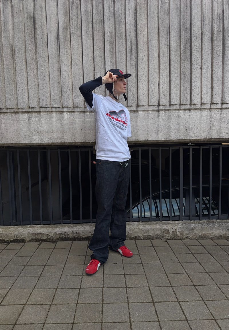 Personne portant un t-shirt blanc à motif, des manches longues noires, une casquette noire, un pantalon large noir et des baskets rouges, debout sur un pavé en carrelage contre un mur en béton.