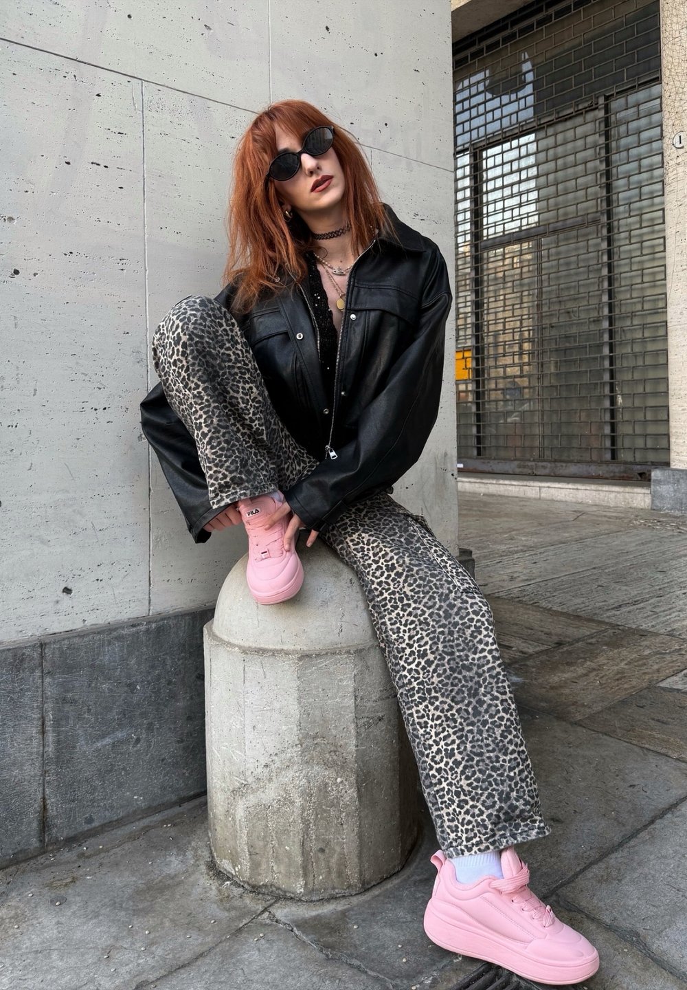 Zwarte leren oversized jas, zwarte top, leopardprint spijkerbroek en roze sneakers. Model zittend op een betonnen paal tegen een gestructureerde muur.