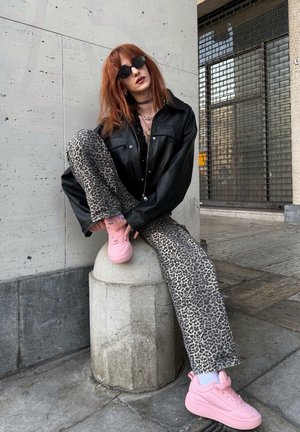 Chaqueta de cuero negra de gran tamaño, top negro, jeans de estampado de leopardo y zapatillas rosas. Modelo sentado en un poste de concreto frente a una pared texturizada.