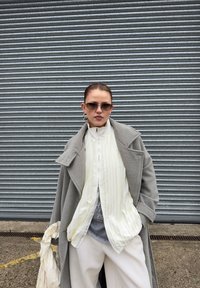 Long manteau gris par-dessus une chemise blanche plissée, t-shirt graphique gris et pantalons blancs amples. Accessoires comprenant des lunettes de soleil et de grandes boucles d'oreilles.