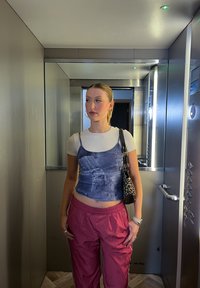 Νεαρή γυναίκα με κότσο και κρίκους στα αυτιά στέκεται μέσα σε έναν μεταλλικό ανελκυστήρα, φορώντας αμάνικο τζιν τοπ, λευκό μπλουζάκι και κόκκινο παντελόνι.