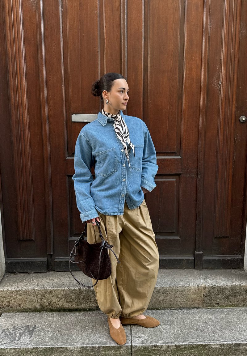 Femme debout devant une porte en bois, portant une veste en jean, un pantalon ample beige, des ballerines marron, une écharpe à imprimé zèbre, et tenant un sac à main noir.