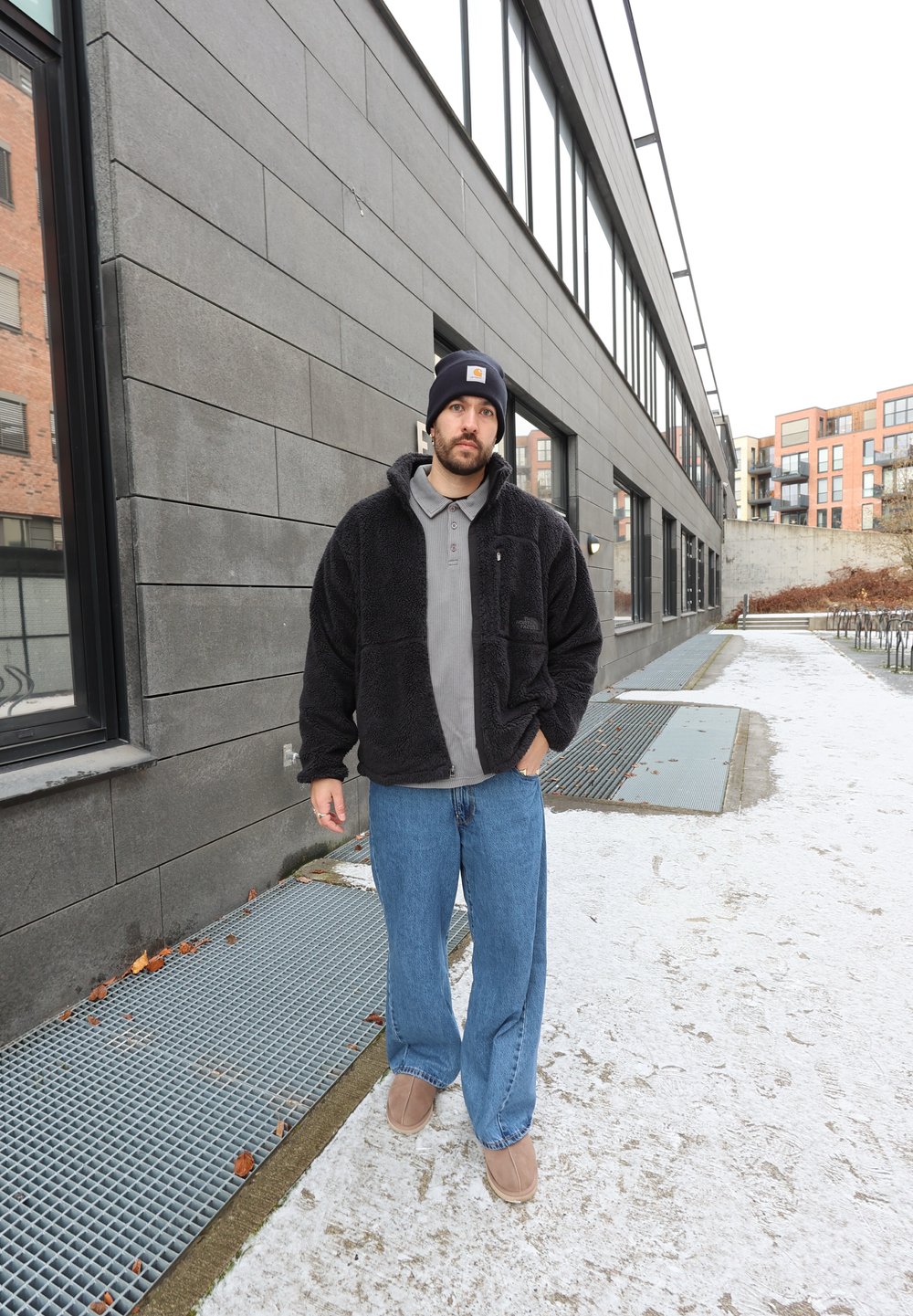 Mees, kes kannab musta fliisjakki, halli polot, siniseid teksaseid ja musta nokamütsi, seisab linnakõnniteel, mis on kaetud lumevärskendusega, halli hoone kõrval.