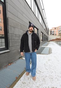 Mees, kes kannab musta fliisjakki, halli polot, siniseid teksaseid ja musta nokamütsi, seisab linnakõnniteel, mis on kaetud lumevärskendusega, halli hoone kõrval.