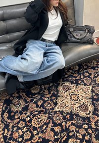 Prevelike svetlo modre jeans hlače z raztrganimi robovi, kombinirane z belo srajco in črnim plaščem, počivajo na sivi usnjeni sedežni garnituri z vzorčastim preprogom.