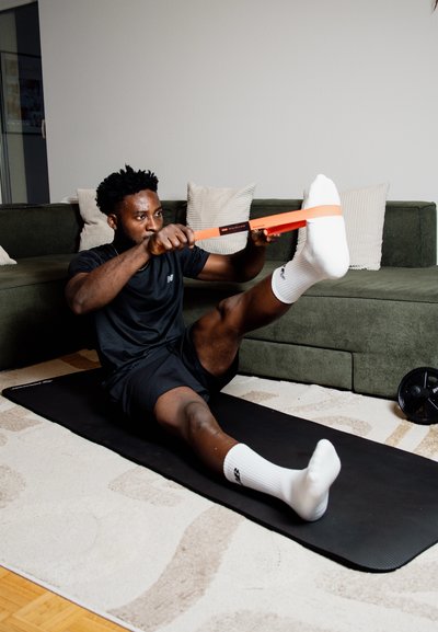 Una esterilla de ejercicio negra sobre una alfombra clara. Una persona vestida con ropa deportiva negra utiliza una banda de resistencia naranja, estirando una pierna hacia adelante.