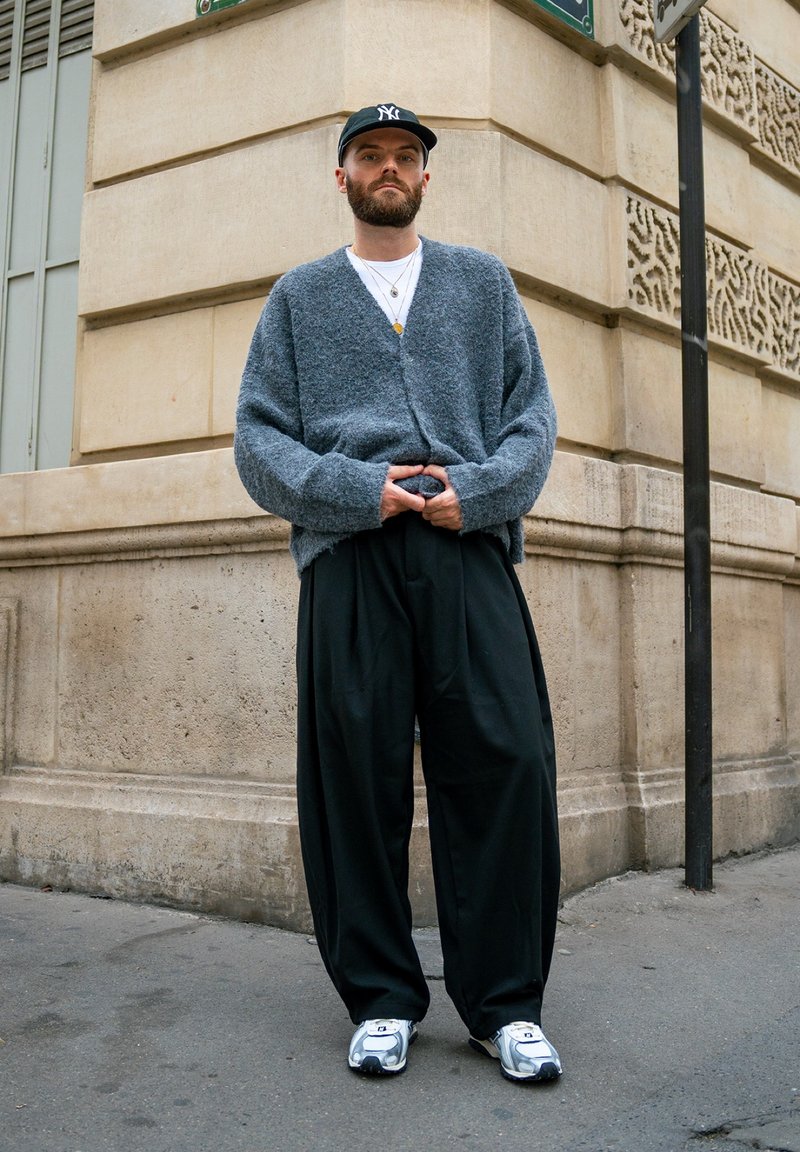 Cardigan gris texturé porté sur une chemise blanche, associé à un pantalon large noir et des baskets argentées métalliques, accessoirisé avec des colliers et une casquette.