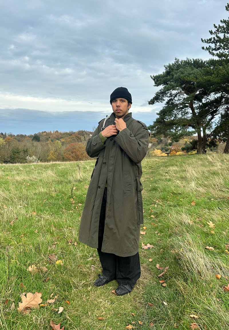 Young person in black beanie and long olive-green coat standing in grassy field with trees and cloudy sky in the background.