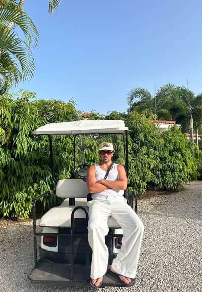 Carrito de golf con chasis de metal negro, toldo blanco, dos asientos y una persona vestida de blanco sentada, rodeada de frondosa vegetación verde.