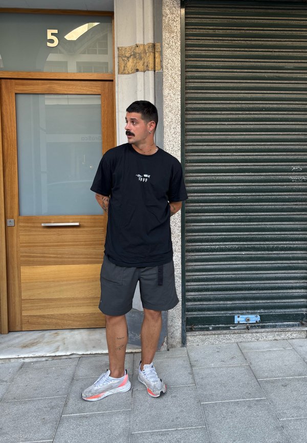 Zwarte t-shirt met witte opdruk, grijze shorts en grijze sneakers. Staat naast een houten deur en een groene metalen luifel. Korte haren en een snor.