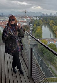 Pledowy poncho z błyszczącym wykończeniem, czarne spodnie i spiczaste botki. Akcesorium: torba na ramię z klamrą. Miejski krajobraz z zielenią i rzeką.