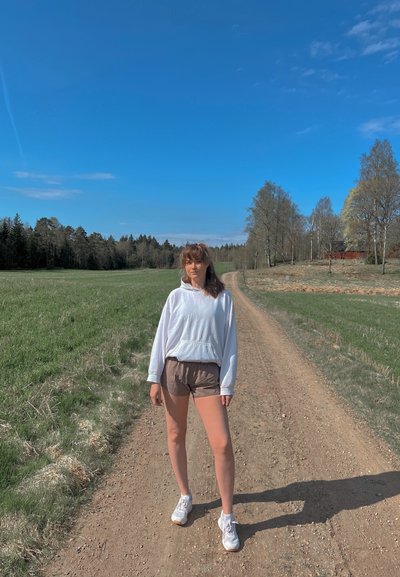 Sudadera blanca con un bolsillo frontal, pantalones cortos marrones y zapatillas blancas. De pie en un camino de tierra rodeado de hierba verde y árboles bajo un cielo azul claro.