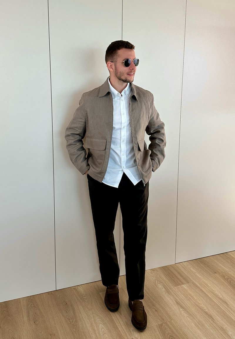 Hombre con gafas de sol, chaqueta beige, camisa blanca, pantalones negros y zapatos marrones, de pie con las manos en los bolsillos frente a una pared blanca.