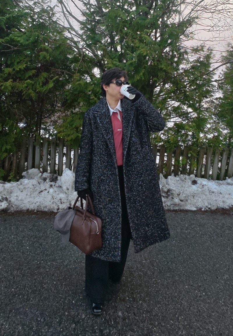 Personne portant un long manteau sombre à motifs et des lunettes de soleil, tenant un sac marron et un gobelet en papier, debout au bord d'une route enneigée avec des arbres en arrière-plan.