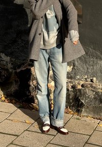 Palton gri din lână, blugi din denim albastru deschis, pulover gri cu grafice și mocasini maro cu accente albe. Cadru exterior cu un zid de piatră.