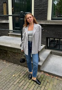 Grau oversized Blazer, graues enges Oberteil, blaue gerade Jeans, schwarze Loafers und eine kleine schwarze Handtasche auf einer Kopfsteinpflasterstraße.