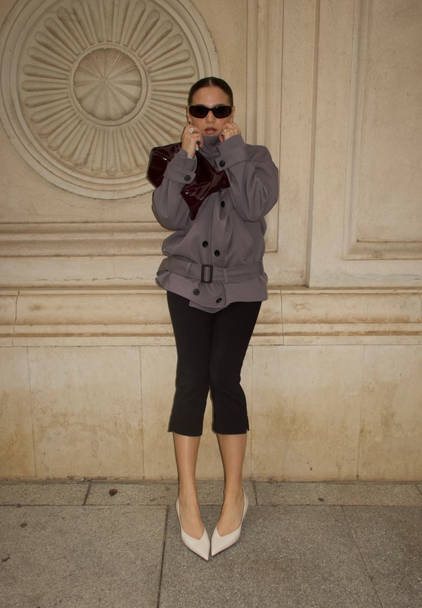 Femme portant une veste grise surdimensionnée, un pantalon court noir, des talons blancs pointus, des lunettes de soleil foncées, tenant une pochette bordeaux foncé contre un mur en pierre.