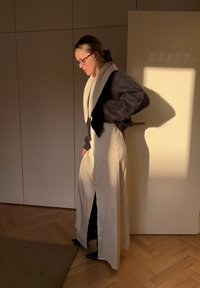 Lang cremefarvet kjole med sidelommer, lagdelt over et sort tøjstykke. Tekstureret mørk sweater med hvid krave, båret med spidse sorte sko.