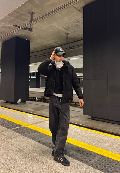 Chaqueta de plumas negra sobre un suéter negro, un cuello alto blanco y unos vaqueros negros holgados. Lleva una gorra verde y gafas de sol, de pie en una plataforma de tren.
