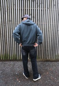 Sweat-shirt gris à capuche avec une coupe décontractée, porté sur un jean foncé et assorti à des baskets noires et blanches, debout contre des lattes en bois.