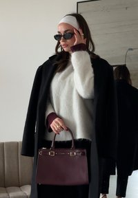 Femme portant un manteau noir sur un pull blanc, tenant un sac  à main violet foncé, ajustant des lunettes de soleil noires, portant un bandeau blanc et du vernis à ongles rouge.