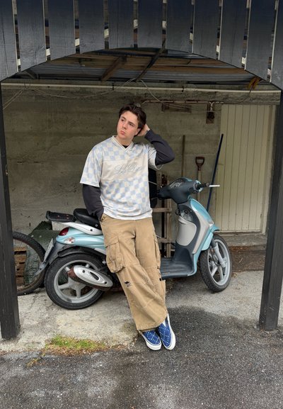 Persona joven apoyada en un scooter azul claro bajo un refugio de madera, con camisa de cuadros, pantalones cargo y zapatillas azules.