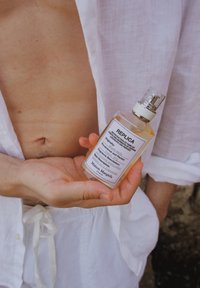 Replika butelki perfum z przezroczystą cieczą, prostokątny kształt, srebrna końcówka rozpylająca i biała etykieta z czarnym tekstem. Trzymana w ręce na tle jasnej tkaniny.