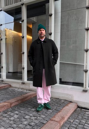 Hombre con un gorro verde, abrigo negro, pantalones rosas y zapatos verdes, de pie sobre escalones de adoquines frente a un edificio moderno con grandes ventanas.