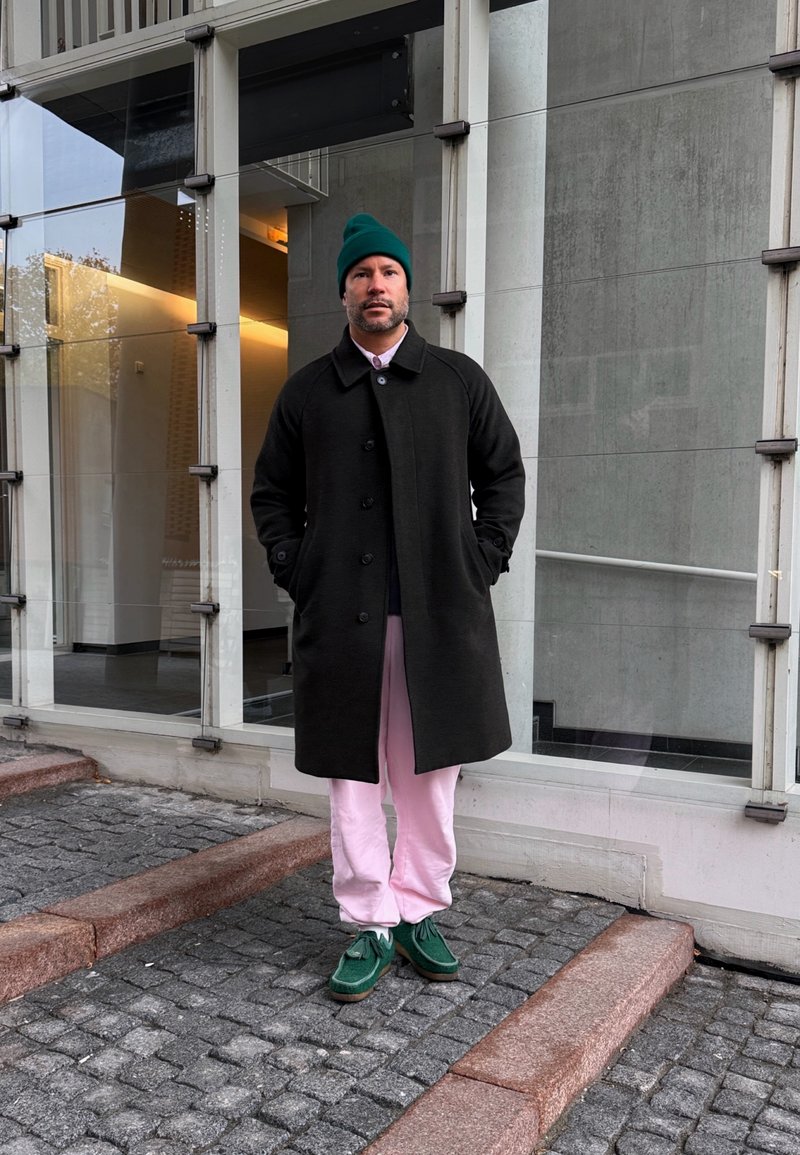 Muž v zelenej čiapke, čiernom kabáte, ružových nohaviciach a zelených topánkach stojaci na kamenných schodoch pred modernou budovou s veľkými oknami.