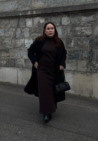 Svart kappa över en mörkbrun polotröja och lång kjol, kombinerat med svarta klackstövlar och en liten handväska, mot en stenvägg.