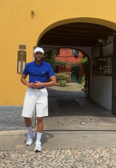 Camisa polo azul, pantalones cortos blancos y gorra blanca, de pie en un arco con un camino de adoquines y vegetación al fondo.