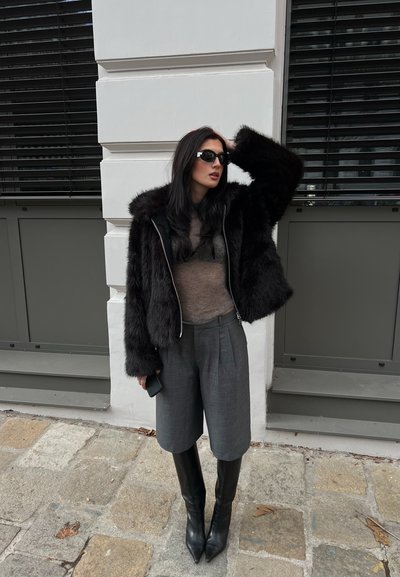 Chaqueta de piel sintética negra con capucha, top gris texturizado, shorts grises plisados y botas negras puntiagudas hasta la rodilla. Gafas de sol y un teléfono negro en la mano.