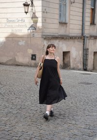 Abito maxi nero con spalline sottili, borsa di paglia intrecciata e calzini bianchi con bordi rossi. Strada acciottolata e edificio consunto sullo sfondo.
